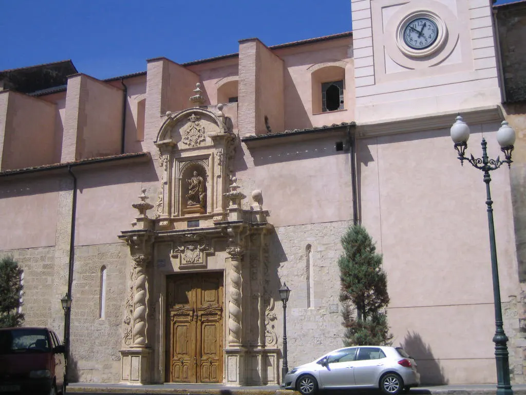 Fachada lateral de la iglesia de Santa Catalina de Alzira: muro de piedra y revoco rosado con una portada barroca de columnas salomónicas y una hornacina con la imagen de la santa; a la derecha, la torre con el reloj y un coche aparcado junto al edificio.