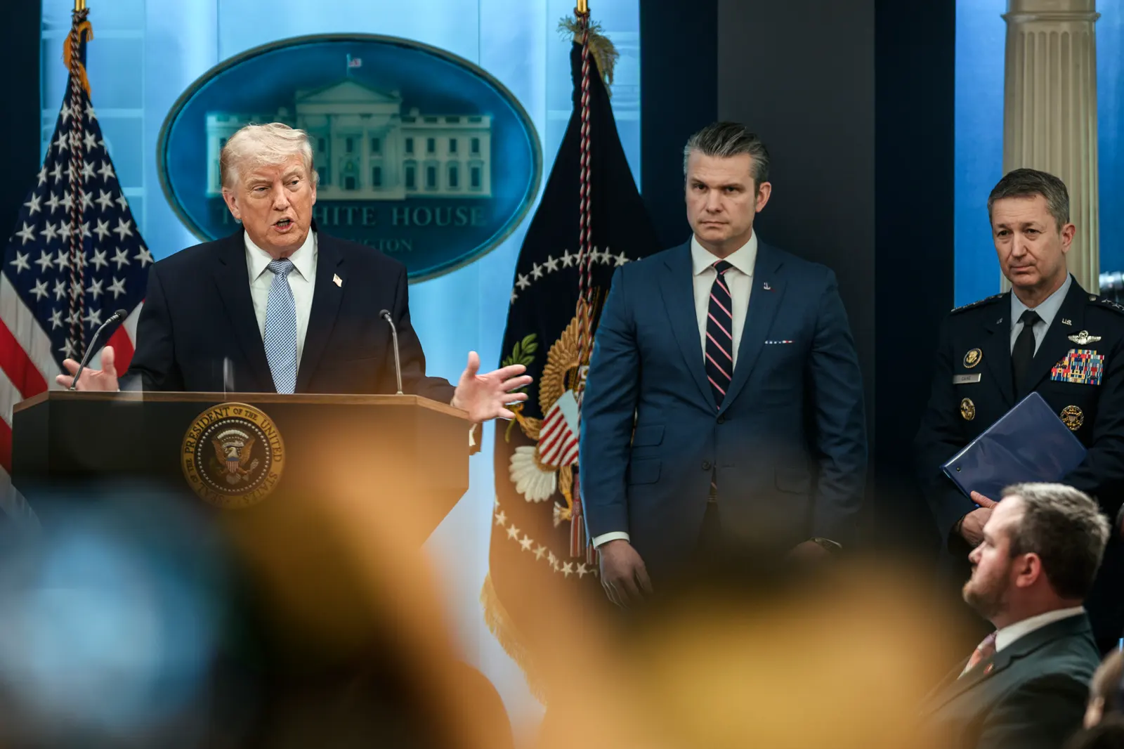 Donald Trump habla con las manos abiertas tras un atril con el sello presidencial en la sala de prensa de la Casa Blanca, entre banderas de Estados Unidos; a su derecha, dos hombres escuchan de pie, uno de traje y otro con uniforme militar de gala.