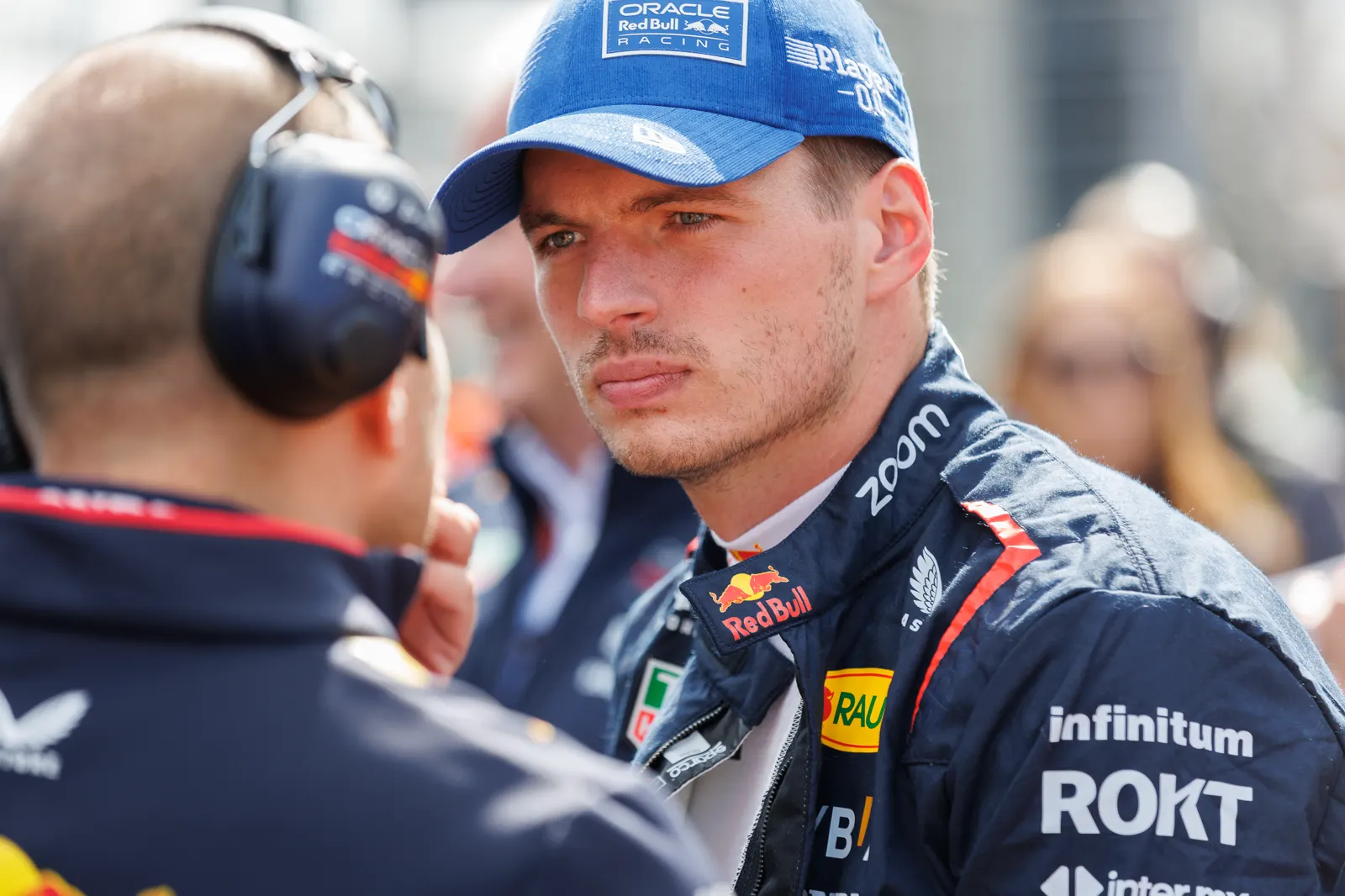 Max Verstappen, con gorra azul y mono de Oracle Red Bull Racing, escucha a un miembro del equipo que lleva auriculares de radio, en la parrilla de salida de un circuito.