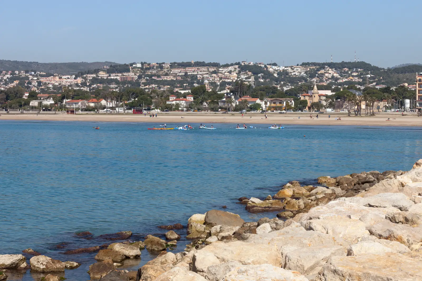 Vista de la playa y el núcleo urbano de Coma-ruga, en El Vendrell, desde un espigón de rocas en primer término; al fondo, casas blancas y ocres entre pinos y una hilera de palmeras junto a la arena, bajo un cielo despejado.