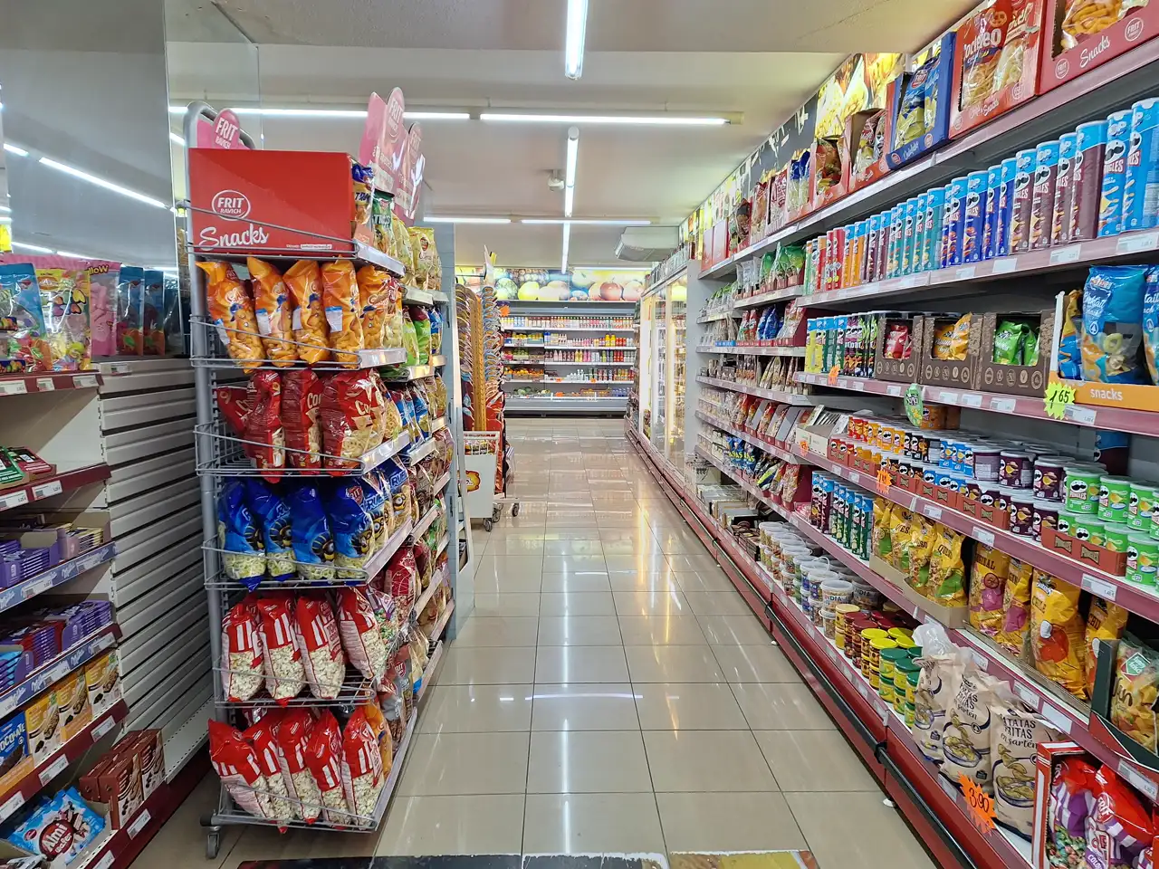 Pasillo de un supermercado con estanterías llenas de bolsas de aperitivos y etiquetas de precios naranjas, sin personas a la vista