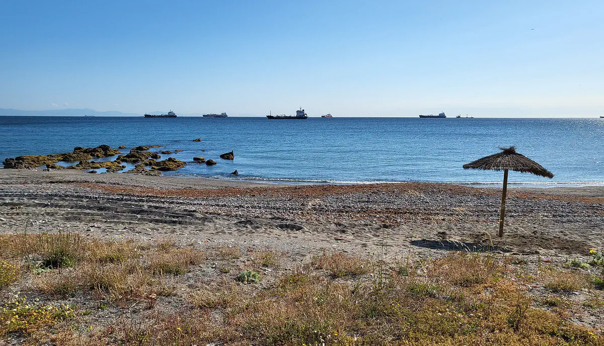 Playa del Trampolín vacía: primer plano de matorral seco y arena con gravilla, una sombrilla de paja solitaria clavada junto a la orilla, el mar en calma y una decena de cargueros fondeados en el horizonte bajo un cielo despejado.