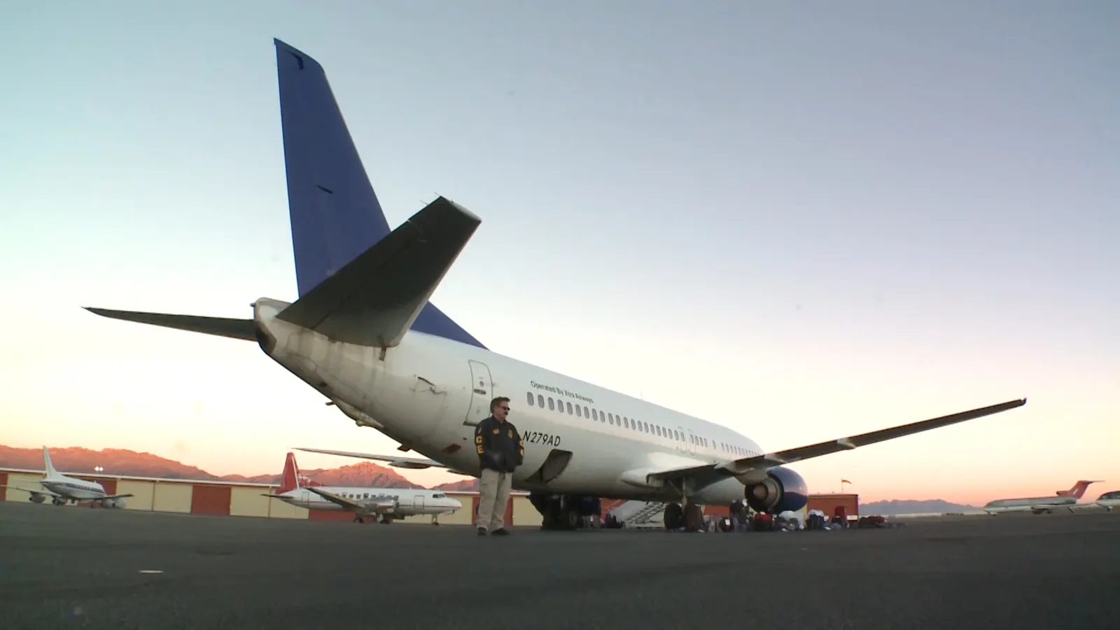 Un agente uniformado con el rótulo ICE en la espalda espera junto a un Boeing 737 blanco estacionado en la pista al amanecer; en el suelo, bajo el ala, se amontonan bolsas y equipajes.