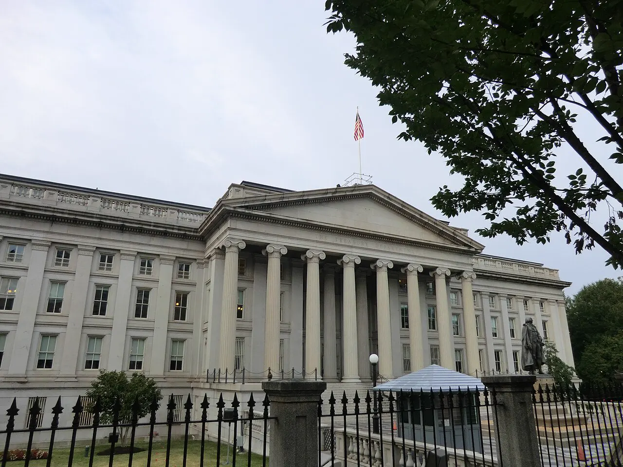 Fachada neoclásica del edificio del Departamento del Tesoro de Estados Unidos en Washington, con su pórtico de columnas y la bandera estadounidense ondeando sobre el tejado, tras una verja de hierro.