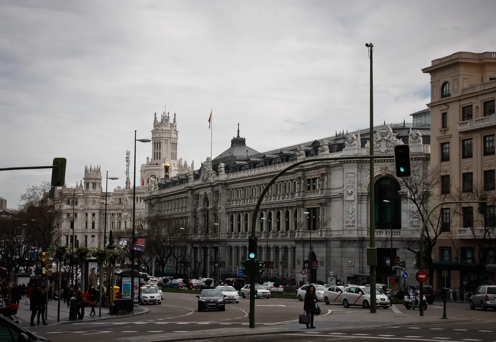 Fachada del edificio del Banco de España en la calle de Alcalá de Madrid, con tráfico y peatones en primer plano bajo un cielo nublado.