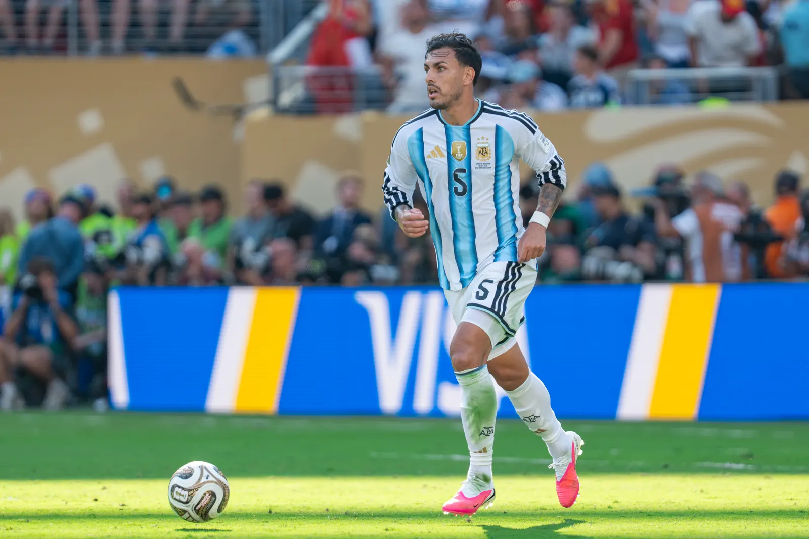 Leandro Paredes, con la camiseta a rayas celestes y blancas de Argentina y el dorsal 5, conduce el balón sobre el césped ante una valla publicitaria y los fotógrafos de banda
