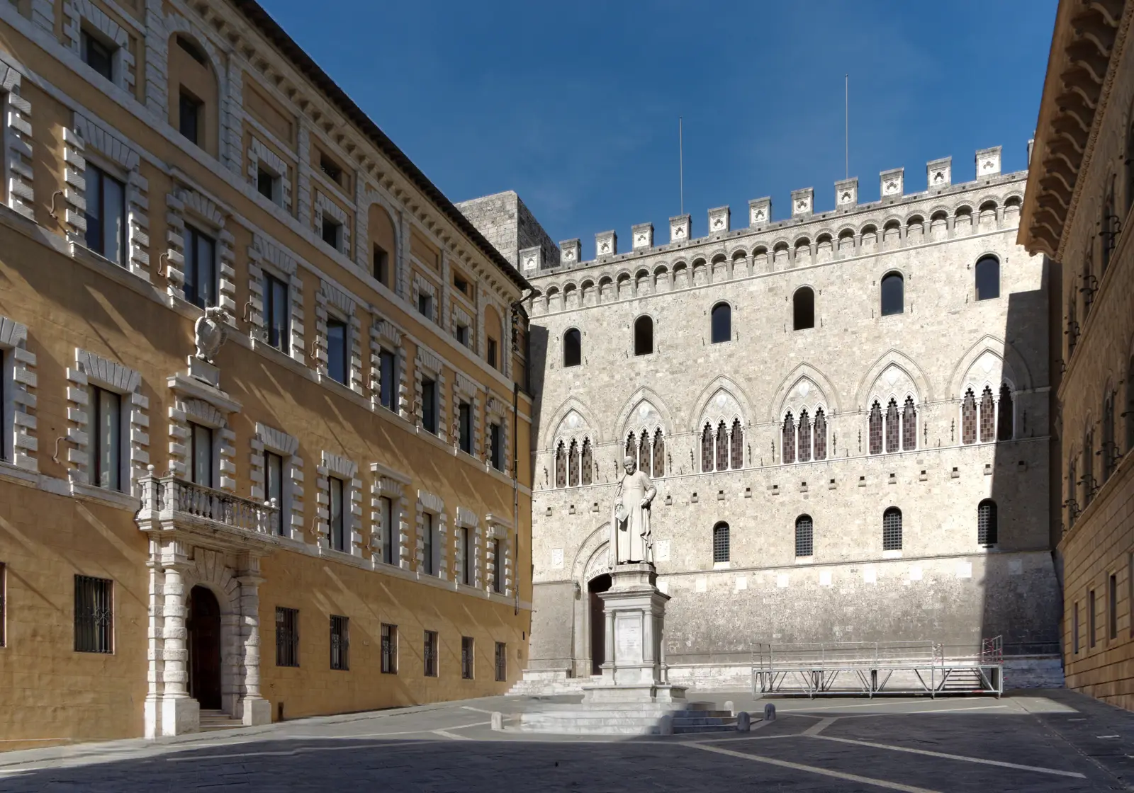 Plaza empedrada de Siena con el Palazzo Salimbeni al fondo, edificio gótico de piedra clara con almenas y ventanas ojivales, sede de Monte dei Paschi; a la izquierda, la fachada amarilla del Palazzo Tantucci, y en el centro una estatua sobre pedestal.