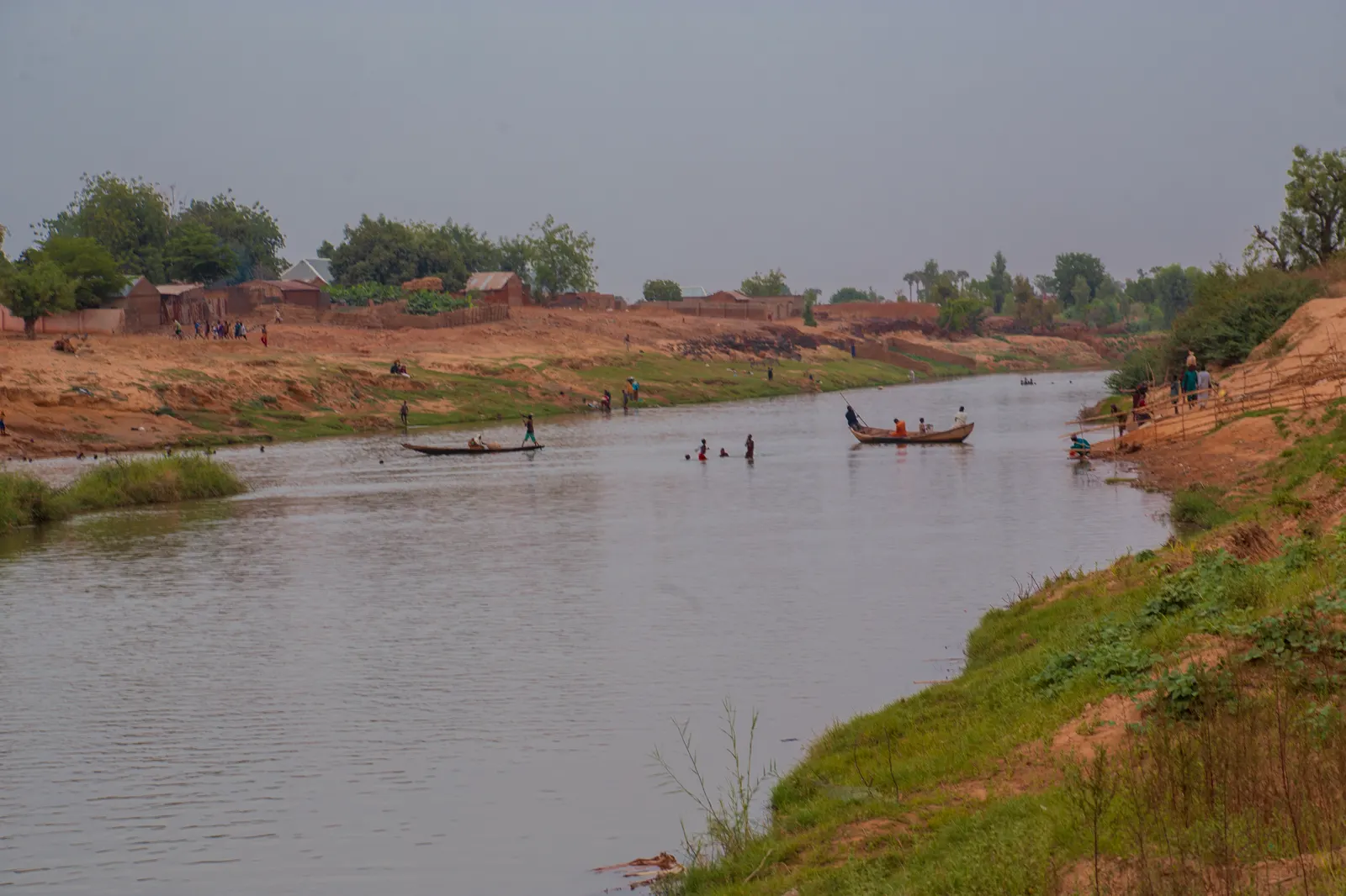 Río ancho y pardo en el noroeste de Nigeria visto desde una orilla de tierra roja. Dos canoas de madera cruzan el cauce con varias personas a bordo y otras vadean el agua; al fondo, un poblado de casas bajas entre árboles bajo un cielo blanquecino.
