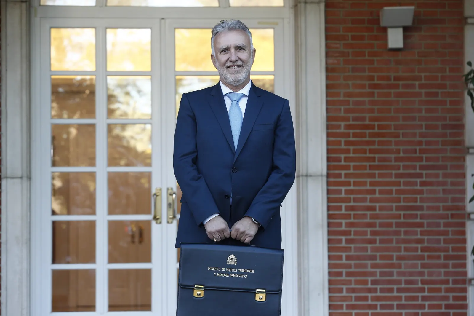 Ángel Víctor Torres, de traje azul marino y corbata celeste, posa sonriente y de pie en la escalinata del edificio del Consejo de Ministros sosteniendo con las dos manos una cartera negra rotulada «Ministro de Política Territorial y Memoria Democrática».