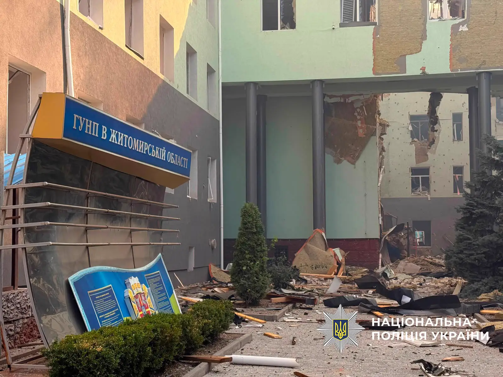 Fachada de un edificio de oficinas con las ventanas reventadas, el revestimiento arrancado y el suelo cubierto de cascotes y tablones; en primer plano, un rótulo azul volcado con el nombre de la jefatura provincial de policía en ucraniano.