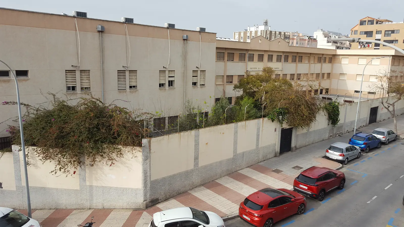 Fachada lateral de un instituto público de tres plantas, de muros claros y ventanas con persianas, tras una tapia encalada cubierta de buganvillas, con coches aparcados en la calle y bloques de viviendas al fondo.