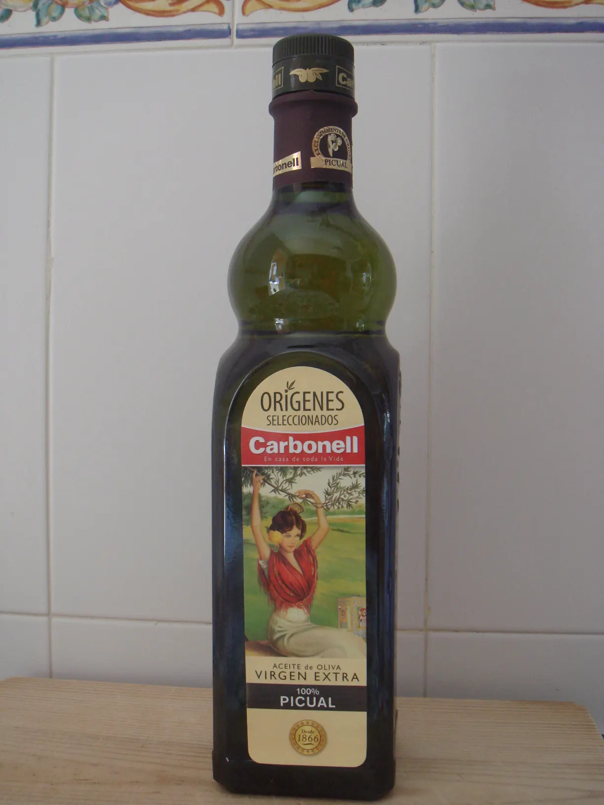 Botella de vidrio oscuro de aceite de oliva virgen extra de la marca Carbonell, con su etiqueta de la bailaora sobre fondo de olivos, apoyada en una encimera frente a una pared de azulejos blancos.