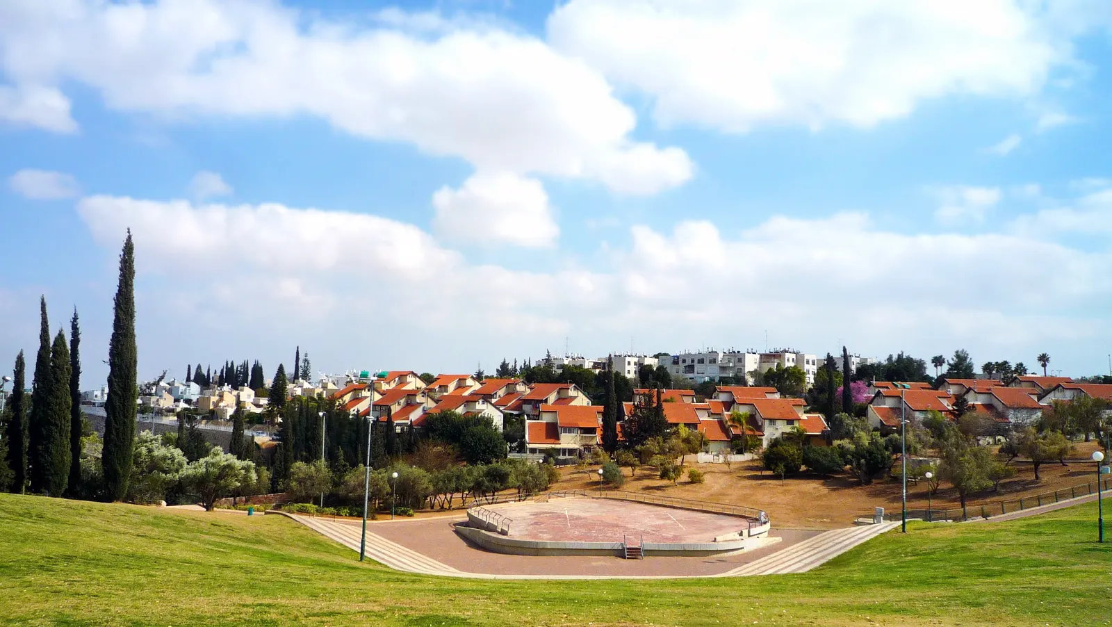 Vista de un barrio de casas bajas con tejados rojos sobre una loma, con cipreses y un parque de césped en primer término, bajo un cielo con nubes.