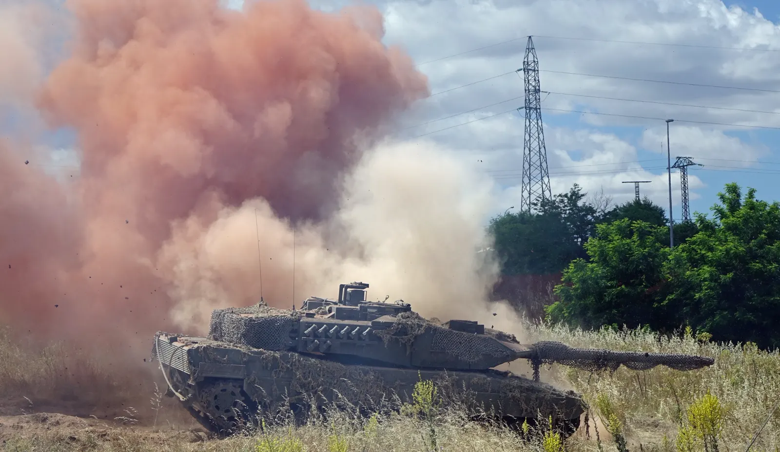 Un carro de combate Leopardo 2E del Ejército de Tierra español, cubierto con red de camuflaje, avanza entre matorrales secos envuelto en una nube de humo naranja y polvo.