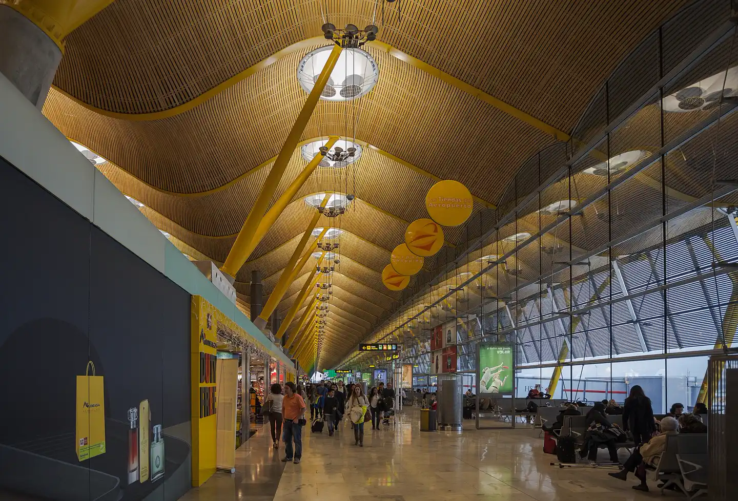Dique de embarque de la Terminal 4 del aeropuerto Adolfo Suárez Madrid-Barajas: techo ondulado de listones de bambú sostenido por pilares amarillos ramificados, tiendas a la izquierda, muro de cristal a la derecha y pasajeros caminando y sentados en la zona de espera.