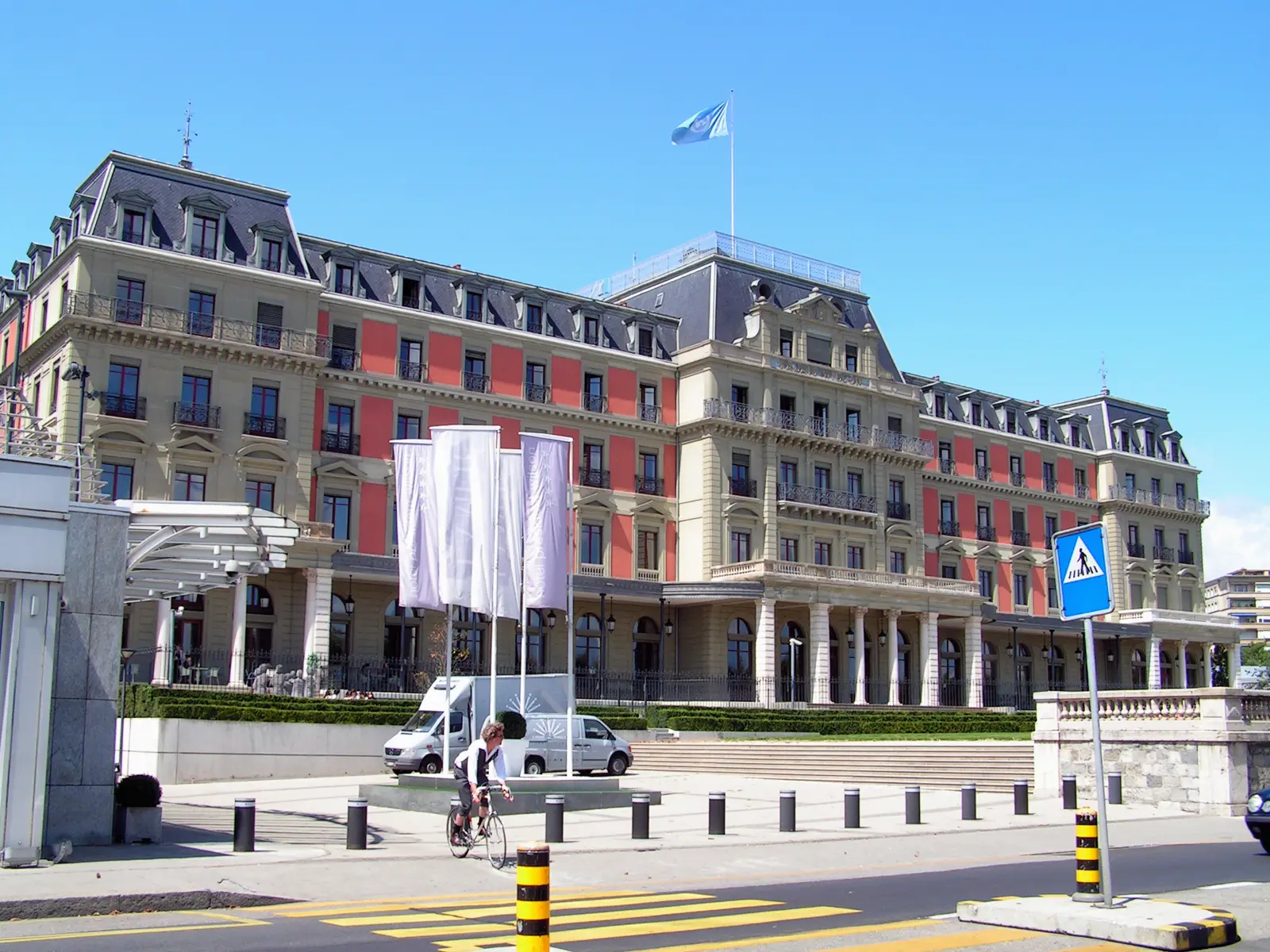 Fachada del Palacio Wilson de Ginebra, un edificio decimonónico de piedra clara y paños rojos con tejado de pizarra, con la bandera de Naciones Unidas ondeando en el tejado y un ciclista cruzando por delante