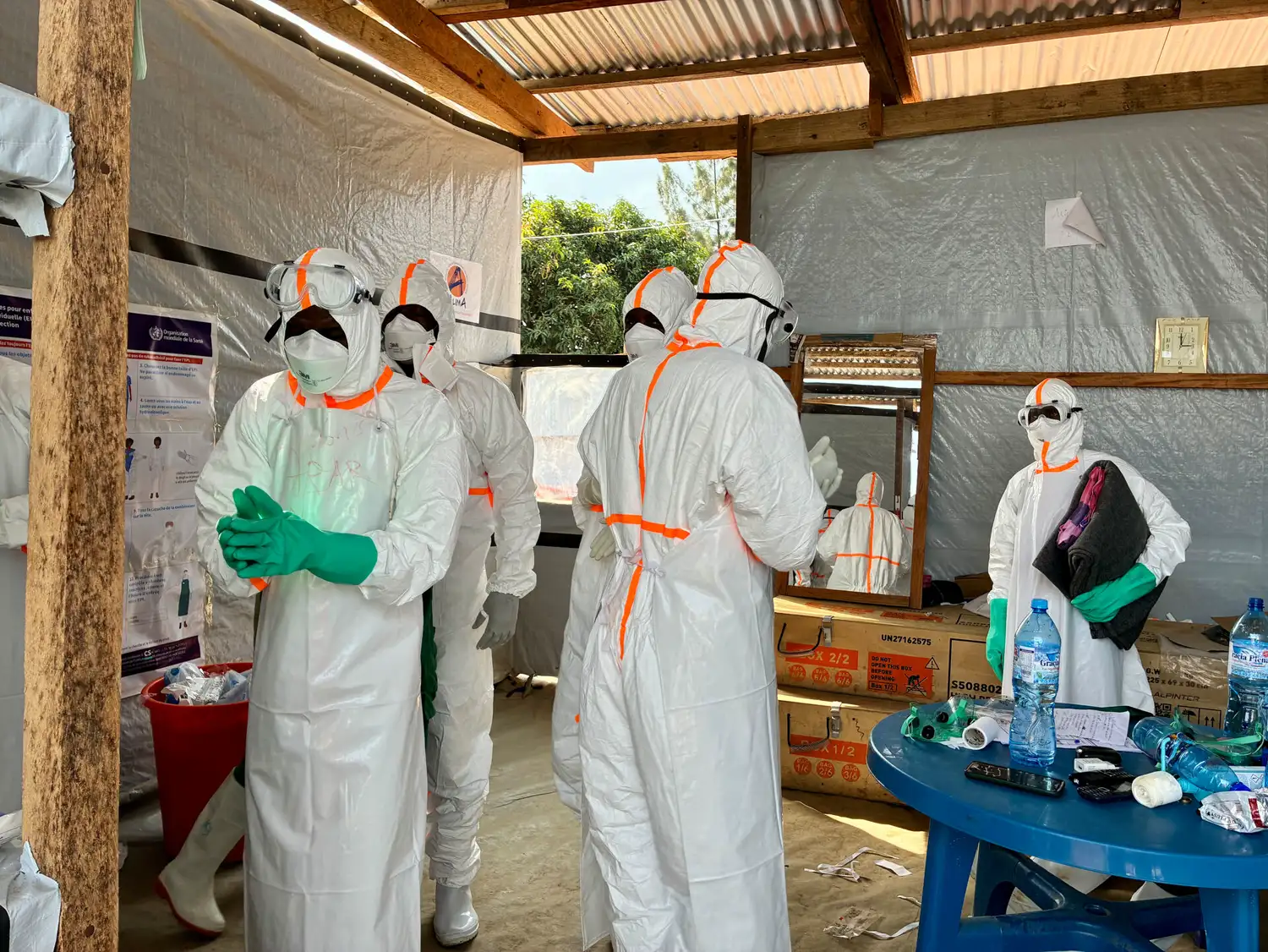 Cinco sanitarios con monos blancos de protección, gafas y mascarilla, uno de ellos con guantes verdes, se ayudan a vestirse dentro de una caseta de lona y chapa con carteles de la OMS en las paredes