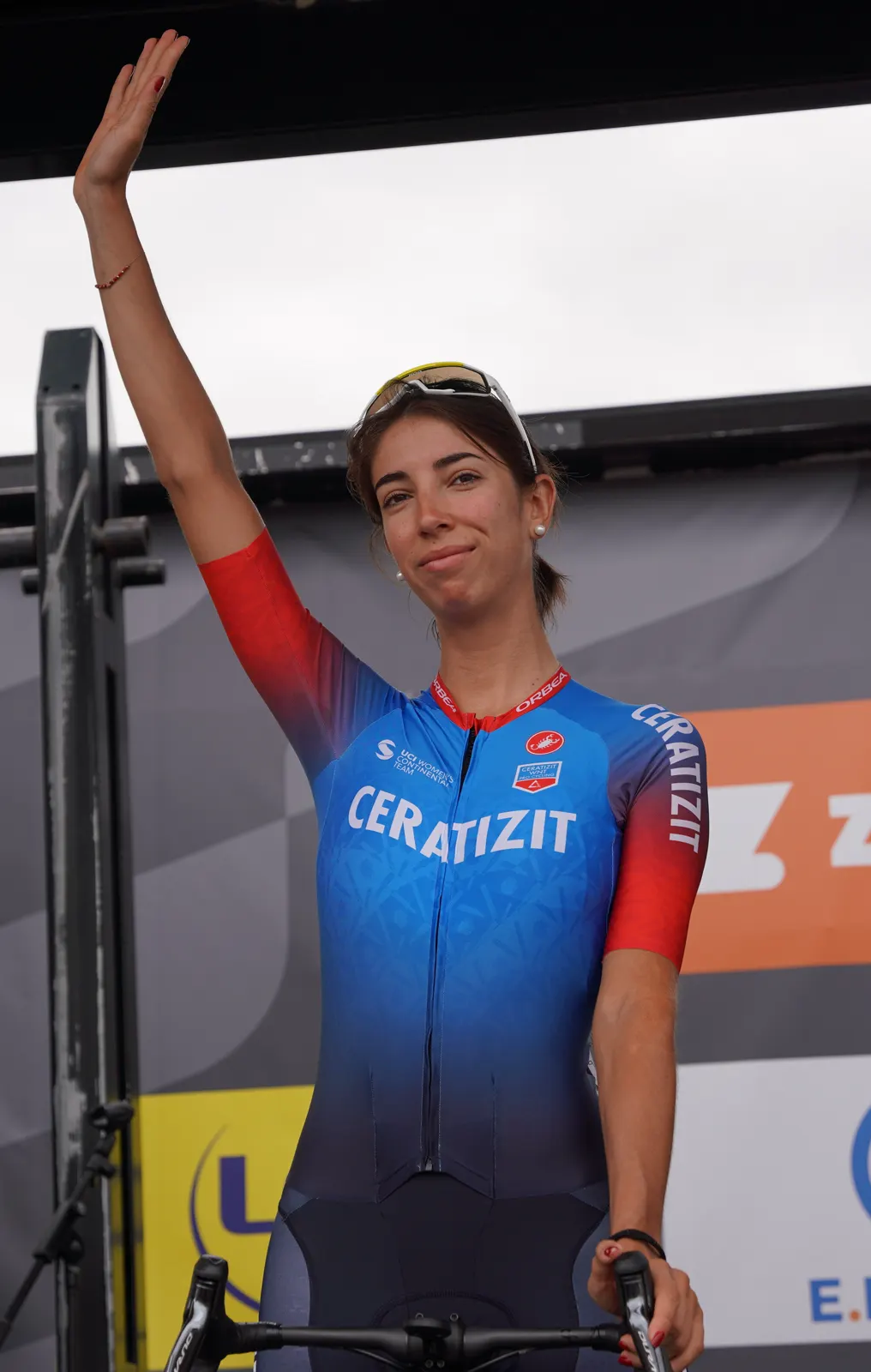 La ciclista Sandra Alonso, con maillot azul y rojo del equipo Ceratizit, saluda con el brazo derecho en alto sobre el escenario de presentación de una carrera.