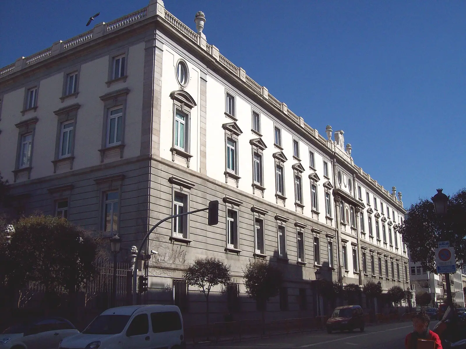 Fachada sur del edificio del Tribunal Supremo en Madrid, vista en escorzo desde la acera: dos plantas de sillería y muro encalado con hileras de ventanas rematadas en frontón, balaustrada y jarrones de piedra en la cornisa, cielo azul despejado y coches aparcados al pie del edificio.