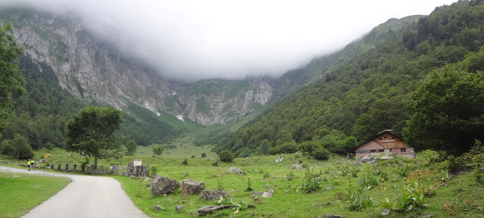 Fondo del circo de la Artiga de Lin, en el Valle de Arán: prados verdes con bloques de roca sueltos, una pista de tierra a la izquierda y una borda de muros de piedra y frontal de madera a la derecha; laderas boscosas a ambos lados y, cerrando el valle, grandes paredes rocosas medio tapadas por un banco de nubes bajas y densas.