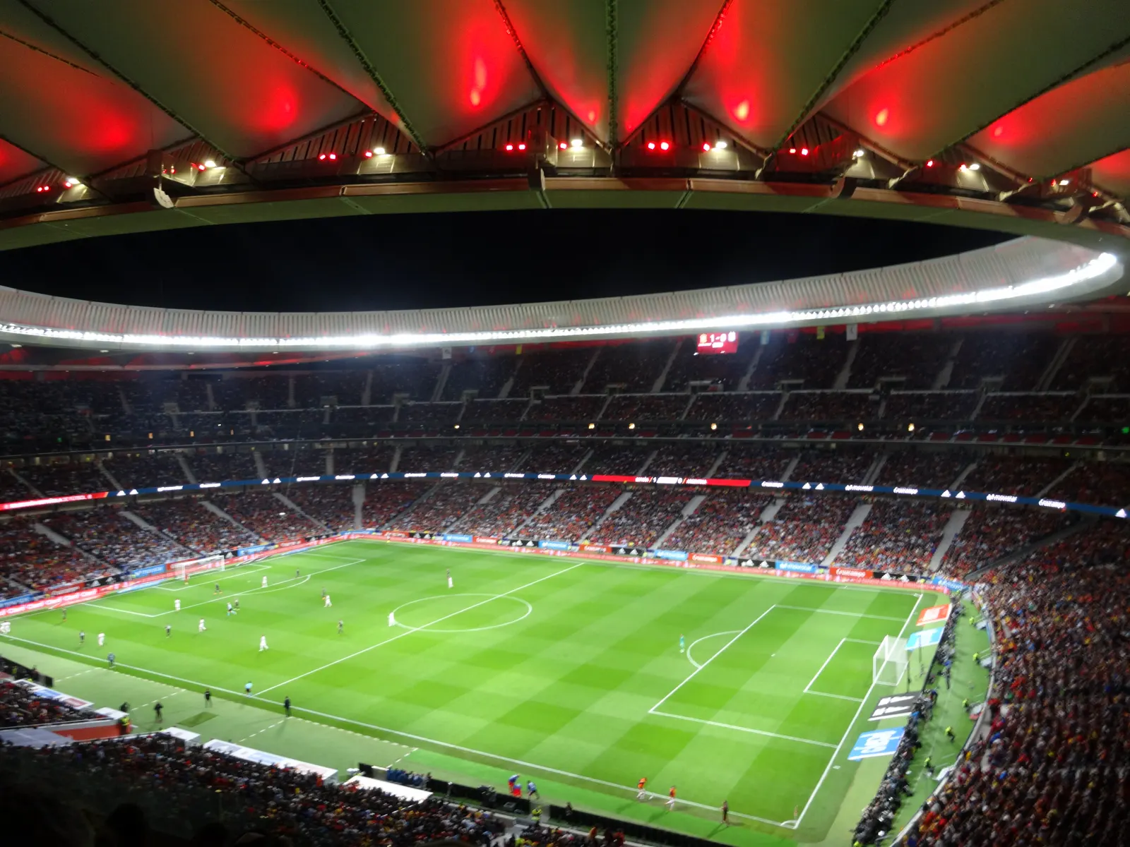 Vista general del estadio Metropolitano de Madrid de noche, con las gradas llenas, el césped iluminado por los focos y la cubierta teñida de rojo.