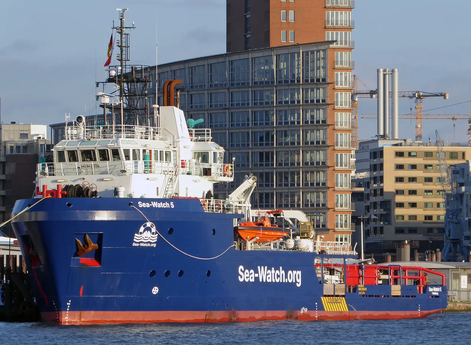 El buque de rescate 'Sea Watch 5', de casco azul con franja roja bajo la línea de flotación, atracado en el puerto de Hamburgo. En el costado se lee «Sea-Watch.org» en letras blancas y en la proa, «Sea-Watch 5»; sobre cubierta hay una lancha de salvamento naranja y, al fondo, edificios de oficinas y grúas de obra.
