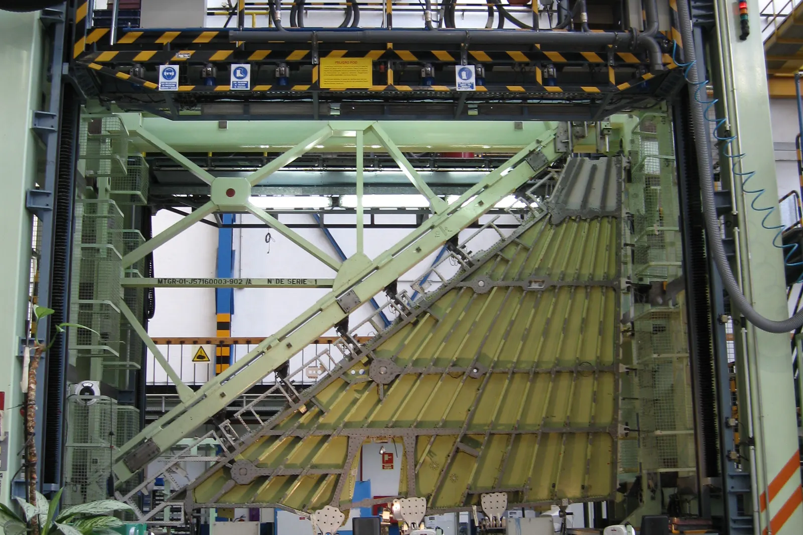 Panel estructural de aeronave, con largueros y remaches a la vista, montado en un utillaje verde de gran tamaño dentro de una nave industrial con pasarelas metálicas y cableado.