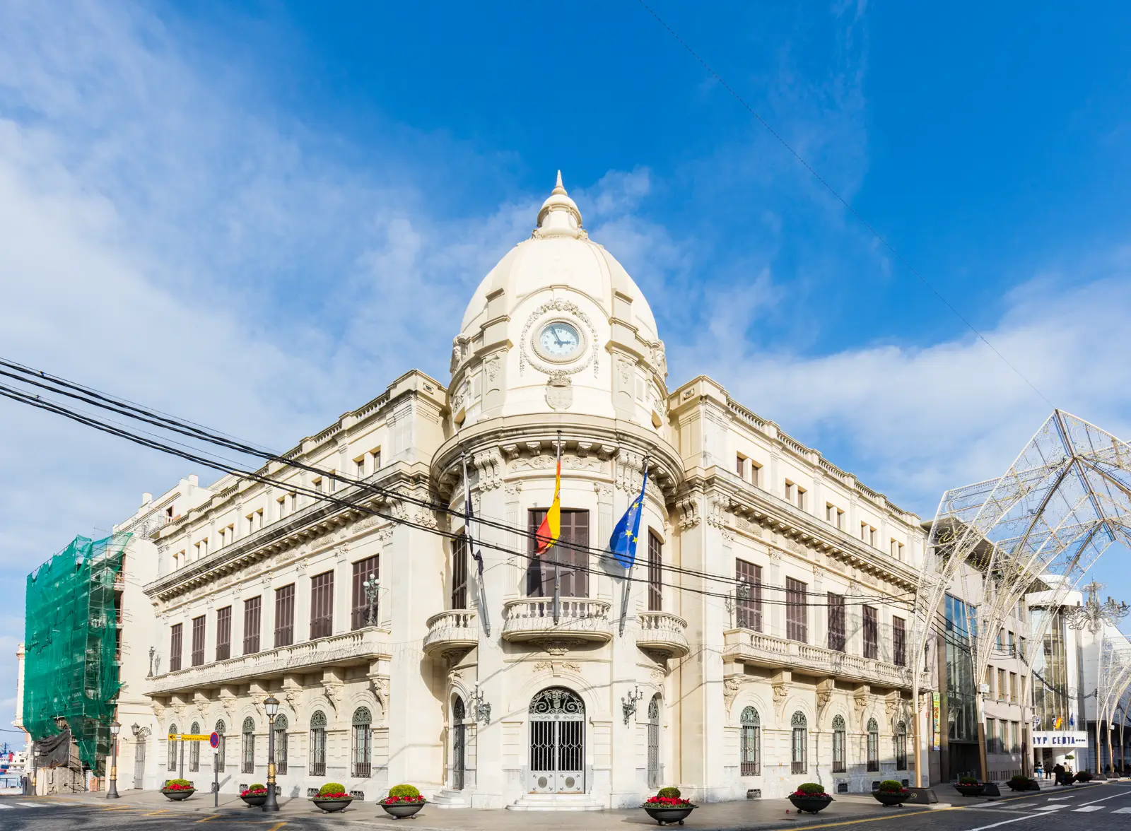 Fachada del Palacio de la Asamblea de Ceuta, edificio blanco de principios del siglo XX con cúpula y reloj sobre el chaflán y las banderas de Ceuta, España y la Unión Europea en el balcón principal, bajo un cielo azul.