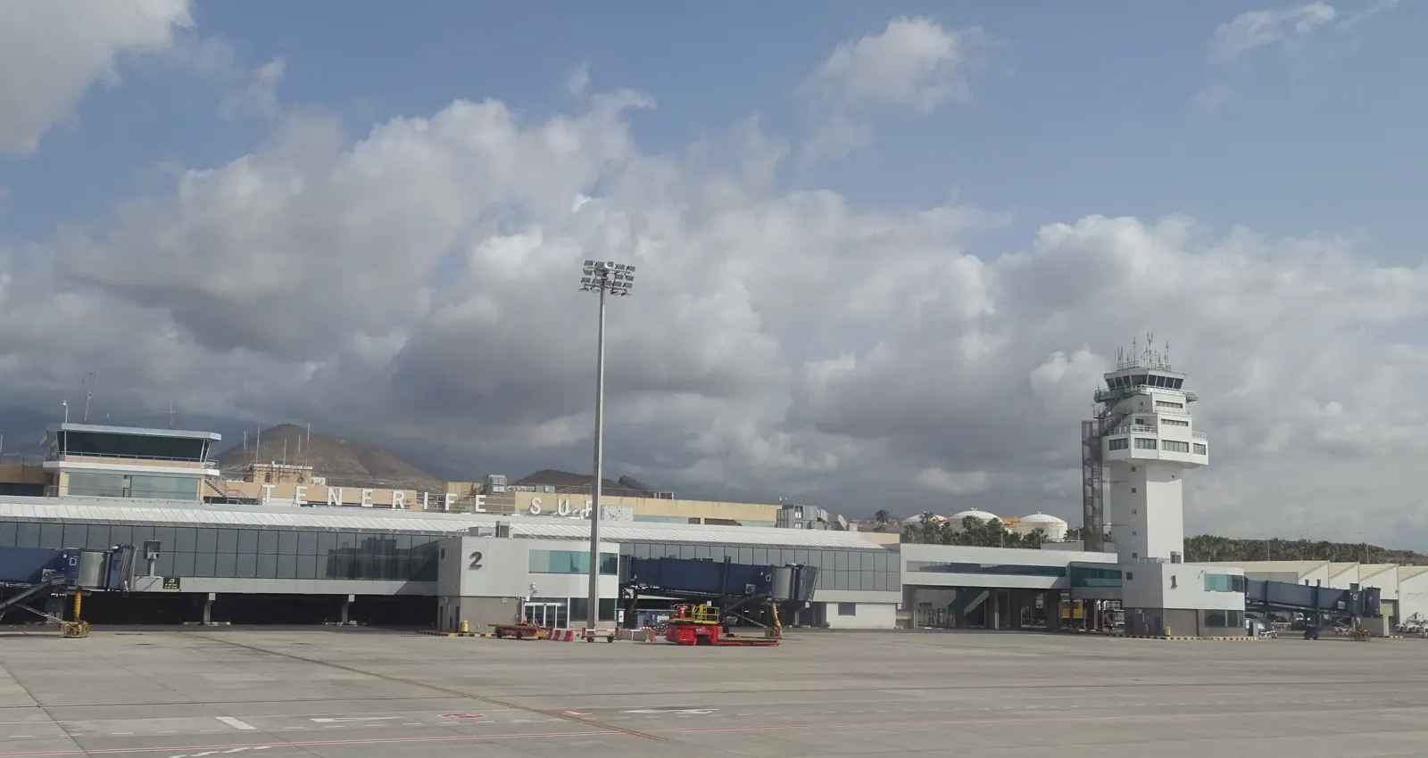 Plataforma del aeropuerto Tenerife Sur vista desde la pista: el edificio terminal con las letras «TENERIFE SUR» en la fachada, varias pasarelas de embarque acristaladas numeradas, vehículos de asistencia en tierra y, a la derecha, la torre de control blanca bajo un cielo nuboso.