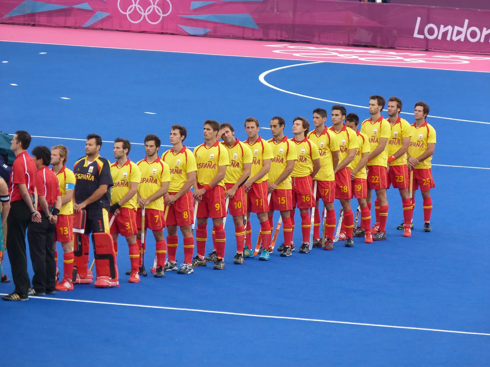 Los jugadores de la selección española masculina de hockey hierba, con camiseta amarilla y pantalón rojo, formados en fila sobre un campo de césped artificial azul antes de un partido.