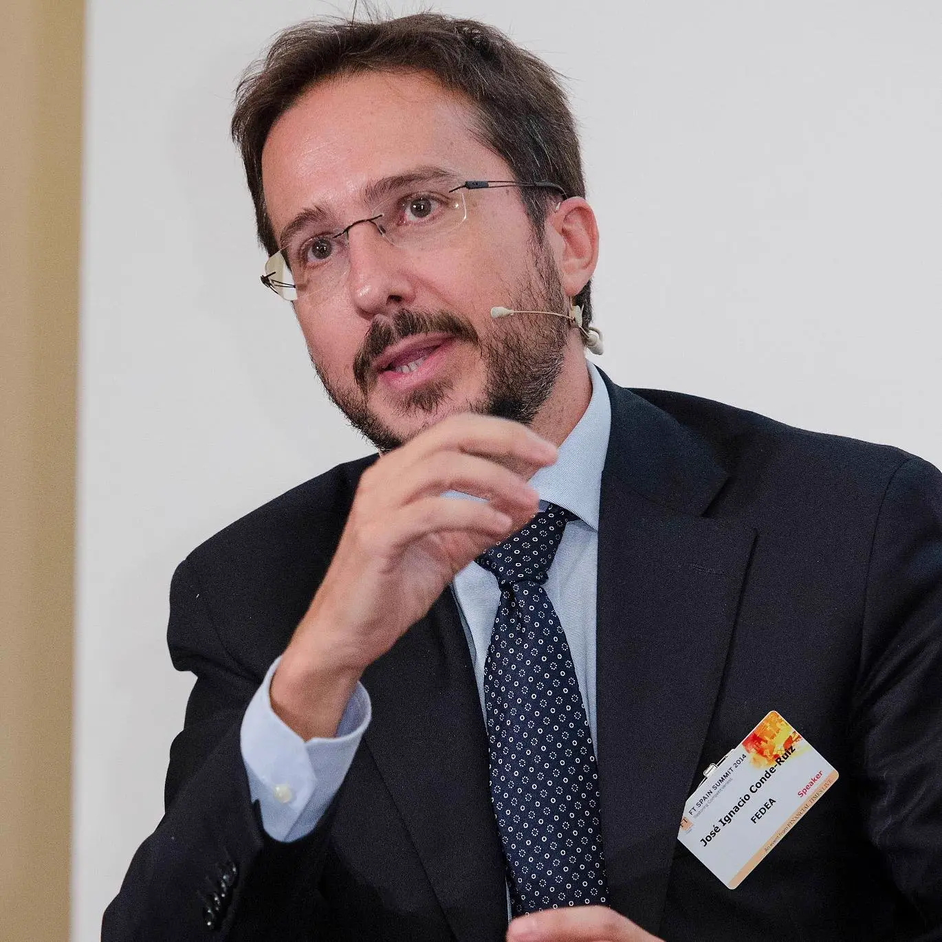 Hombre con gafas y barba, traje oscuro y corbata azul, habla con un micrófono de diadema y gesticula con la mano derecha; lleva prendida una acreditación de ponente.