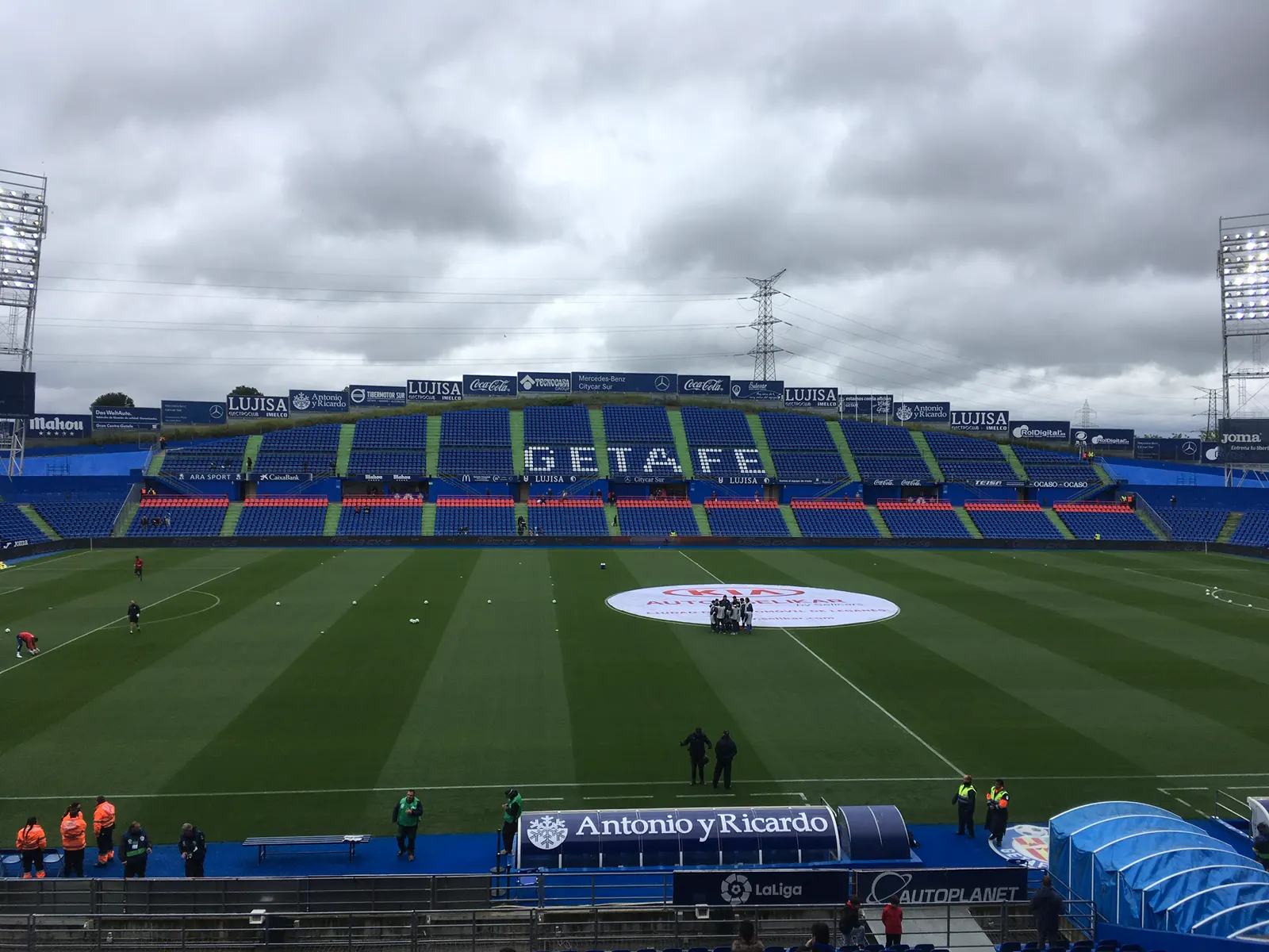 Interior del estadio Coliseum Alfonso Pérez de Getafe antes de un partido, con el césped rayado y vacío, un grupo de jugadores reunido en el círculo central y la grada de fondo con asientos azules que dibujan la palabra «Getafe» bajo un cielo nublado.