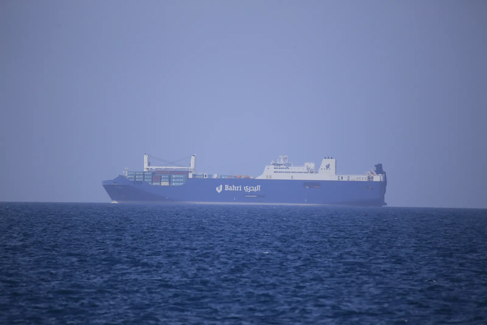 Un buque de casco azul con el logotipo y el nombre 'Bahri' rotulados en el costado navega en mar abierto bajo un cielo brumoso.