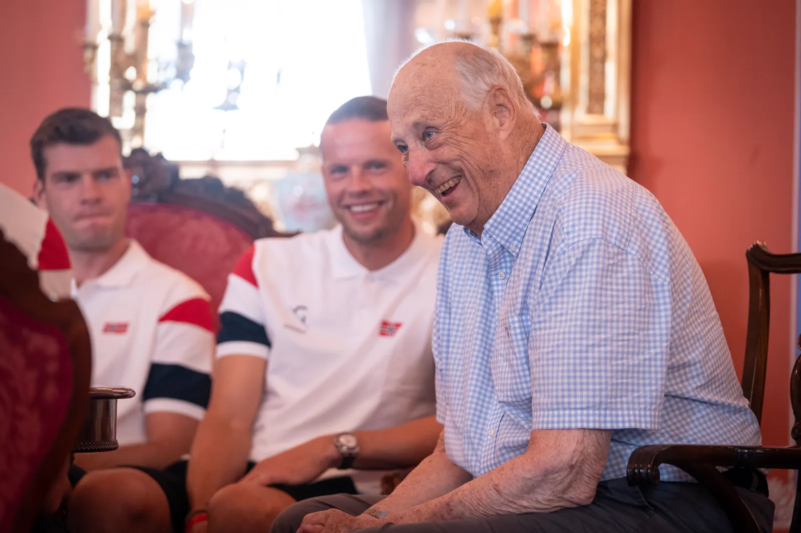 El rey Harald V de Noruega, sentado y sonriente, conversa con varios jugadores de la selección noruega de fútbol en una sala del Palacio Real de Oslo.
