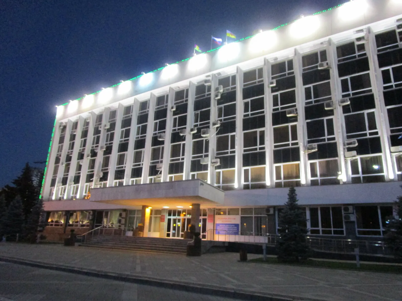 Edificio institucional de varias plantas iluminado de noche, con columnas blancas en la fachada y una bandera rusa ondeando en la cubierta, visto desde la acera de enfrente.