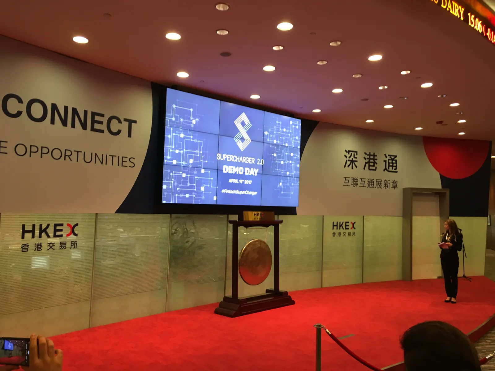 Escenario de ceremonias de la Bolsa de Hong Kong, con un gran gong dorado sobre alfombra roja, el rótulo HKEX en la pared y una pantalla encendida al fondo.