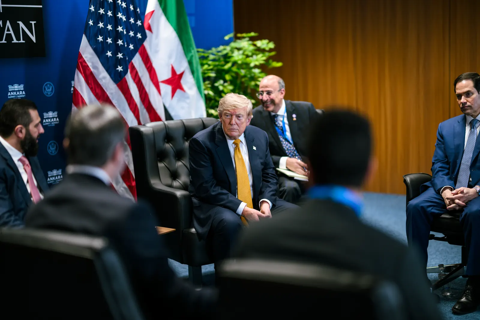 El presidente de Estados Unidos, Donald Trump, sentado en un sillón durante una reunión bilateral, con la bandera estadounidense y la bandera siria de tres estrellas rojas desplegadas a su espalda y varios asesores sentados alrededor.