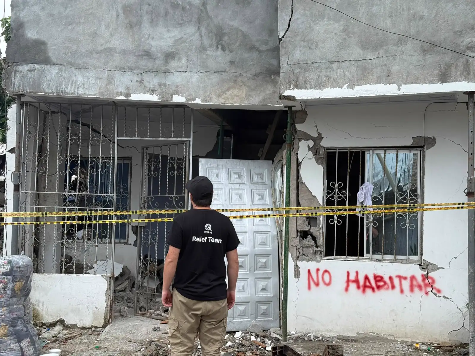 Fachada de una casa de hormigón agrietada de arriba abajo, con la pared reventada junto a la puerta metálica y las ventanas enrejadas rotas. Sobre el muro blanco alguien ha pintado con espray rojo «NO HABITAR». Delante cruza una cinta amarilla de balizamiento con la leyenda «PELIGRO NO PASE» y, de espaldas, un hombre con gorra y camiseta negra de un equipo de ayuda humanitaria mira la vivienda.