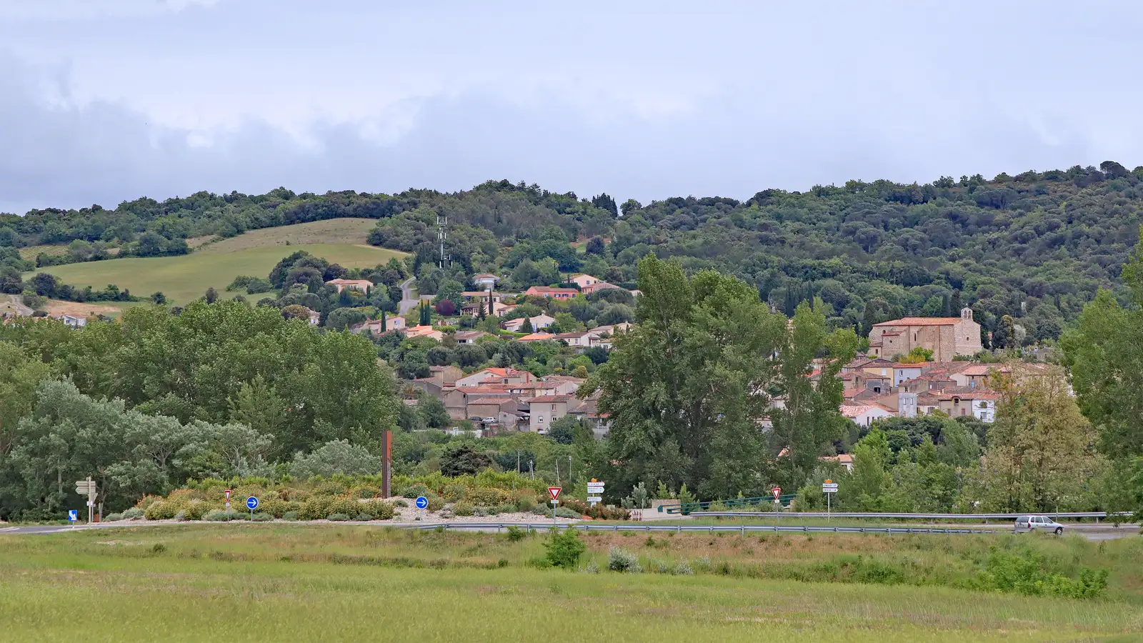 Vista general del pueblo de Pomas desde el norte: casas de tejado de teja roja escalonadas en la ladera, la torre de la iglesia a la derecha, colinas boscosas al fondo y, en primer plano, un prado, una carretera con una rotonda y un coche.