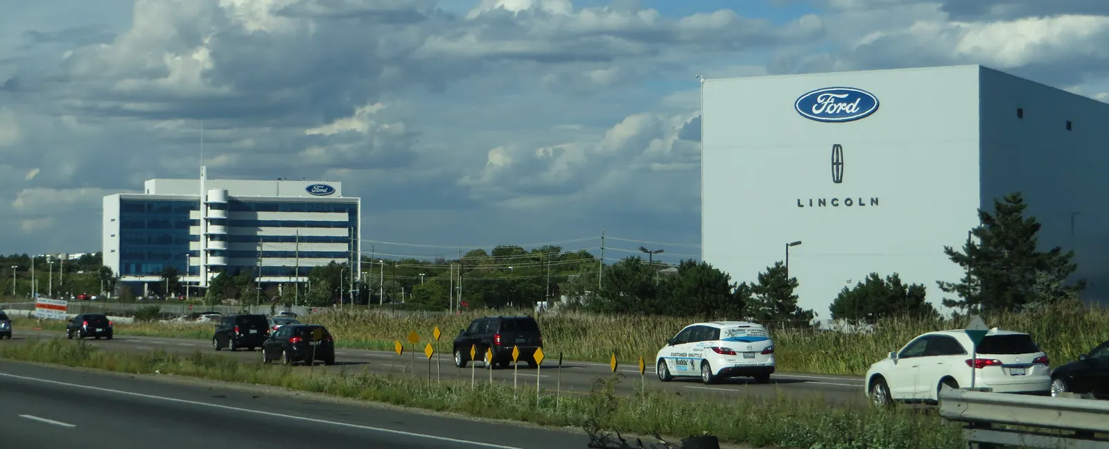 Vista desde la autopista de la planta de montaje de Ford en Oakville (Ontario): una nave blanca con el óvalo azul de Ford y el logotipo de Lincoln, junto a un edificio de oficinas acristalado; en primer plano, coches circulando bajo un cielo nublado.