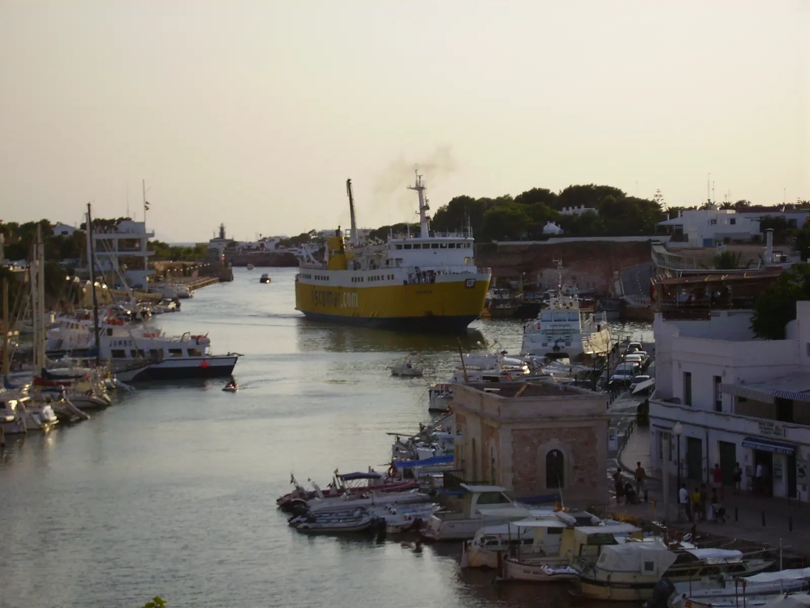 Vista elevada del puerto de Ciutadella de Menorca al atardecer: un canal de agua largo y estrecho entre dos muelles, con decenas de embarcaciones amarradas a ambos lados y un ferry amarillo y blanco navegando hacia la bocana.