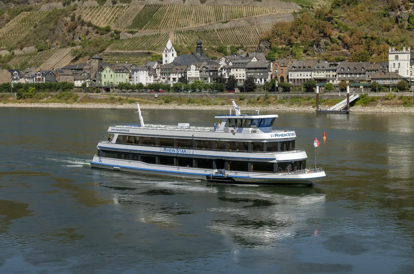 Barco fluvial de pasajeros blanco con el rótulo «Rhein Star» navegando por un Rin de aguas muy bajas, con bancos del lecho asomando bajo la superficie; al fondo, el pueblo de Kaub con su iglesia y laderas de viñedos en terrazas, y a la derecha un embarcadero flotante hundido sobre sus pilotes metálicos.