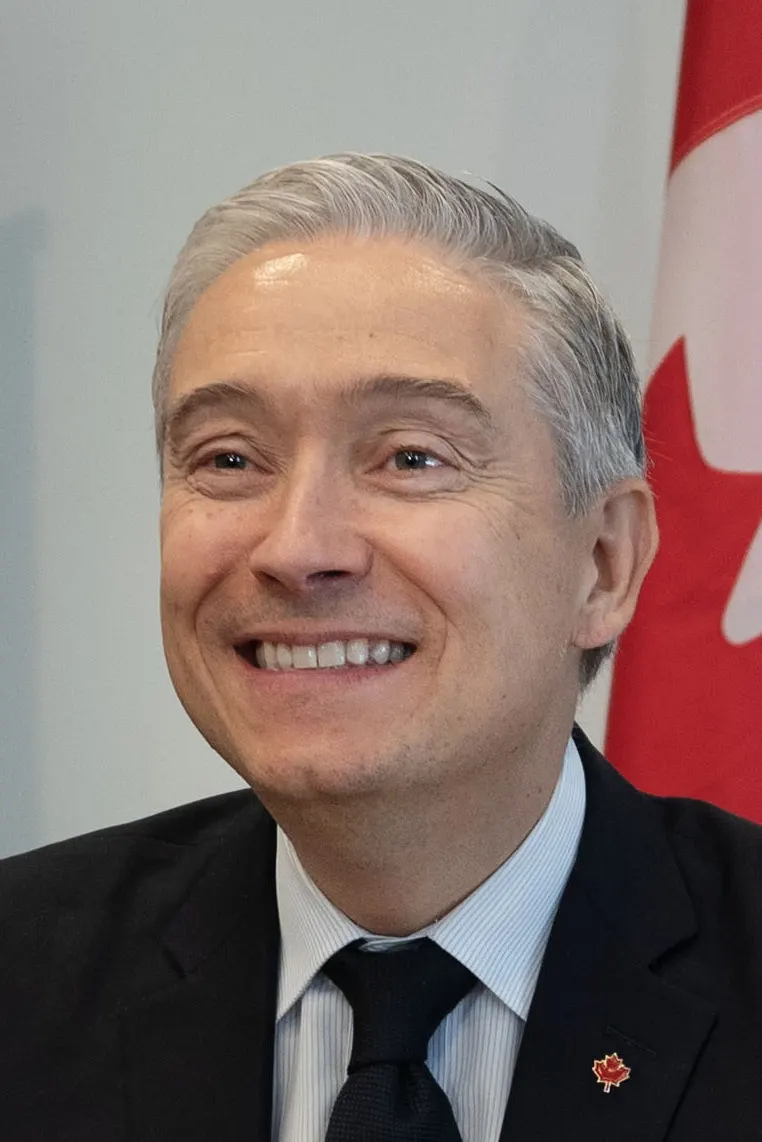 Retrato de François-Philippe Champagne sonriendo, con traje oscuro, camisa de rayas azules y corbata negra, con un pin de la hoja de arce en la solapa; detrás, la bandera de Canadá.