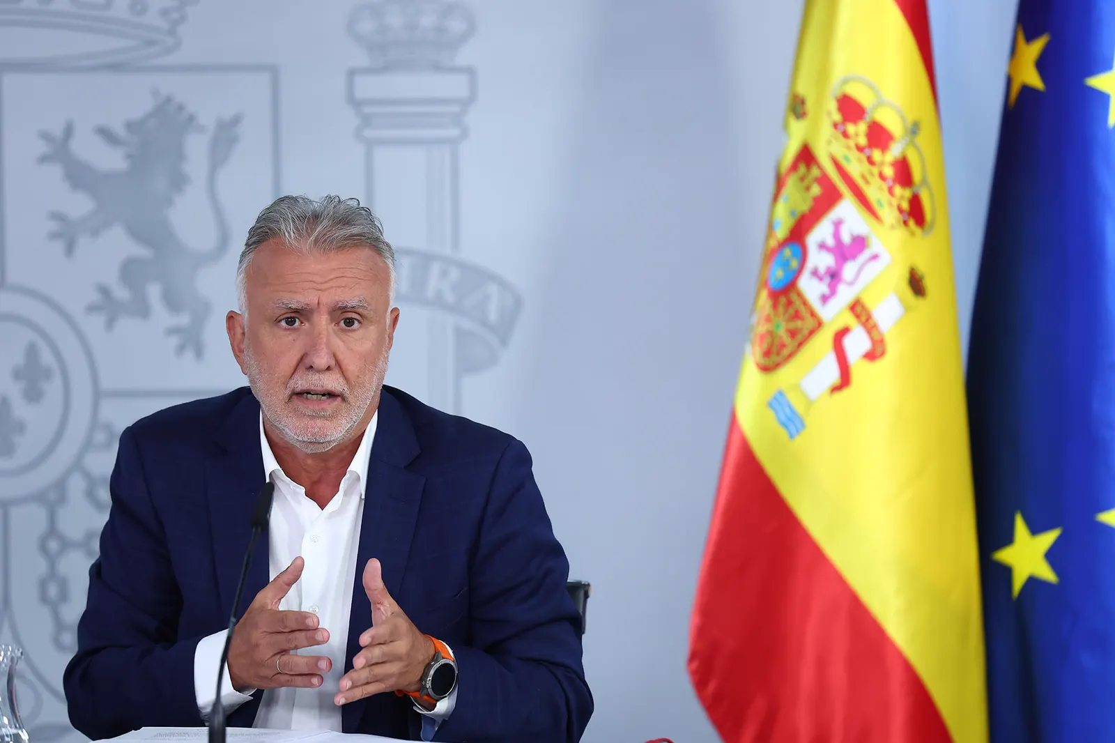 El ministro Ángel Víctor Torres, sentado ante un micrófono y con las banderas de España y de la Unión Europea a su derecha, habla y gesticula con las dos manos durante la rueda de prensa posterior al Consejo de Ministros; al fondo, el escudo de España sobre un panel gris.