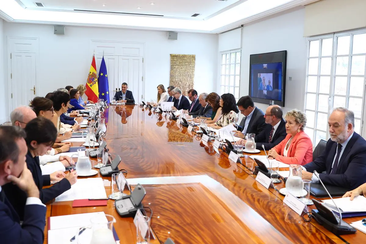 Pedro Sánchez, sentado en la cabecera de una larga mesa de madera brillante y con las banderas de España y de la Unión Europea a su espalda, preside una reunión con una veintena de ministros y altos cargos sentados a ambos lados; en la pared derecha, una pantalla muestra a los asistentes por videoconferencia.