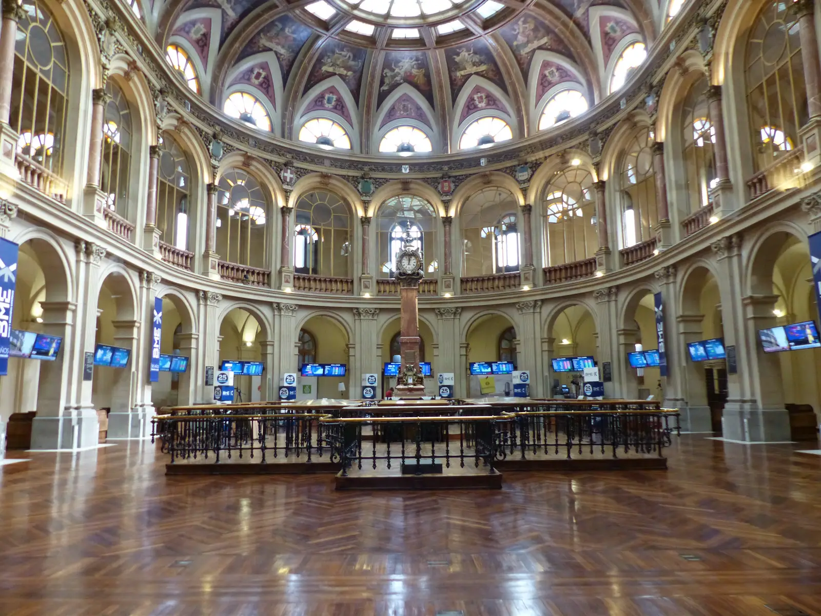 Sala de contratación circular del Palacio de la Bolsa de Madrid, vacía, con el suelo de madera brillante, el corro central de barandillas de hierro y pantallas de cotización encendidas bajo la cúpula pintada.