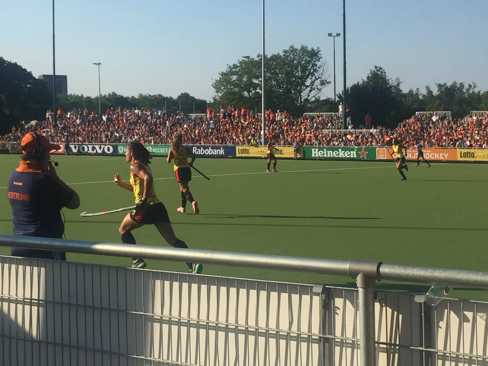 Jugadoras de la selección española femenina de hockey hierba, con camiseta amarilla y falda negra y roja, corren sobre un campo de césped artificial verde durante un partido contra Países Bajos, con la grada llena al fondo.