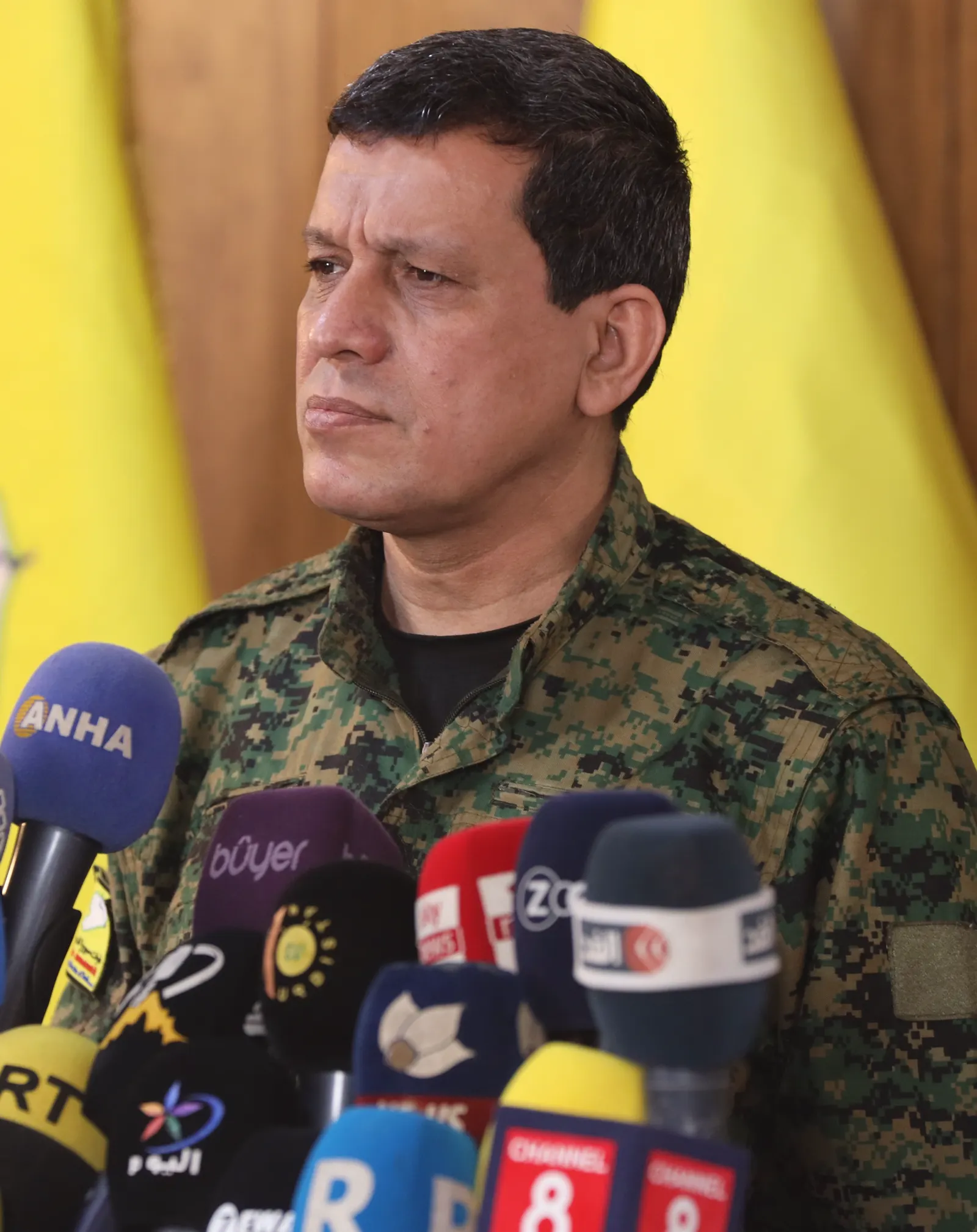 Mazloum Abdi, con uniforme militar de camuflaje, comparece ante un grupo de micrófonos de varias televisiones, con banderas amarillas al fondo.