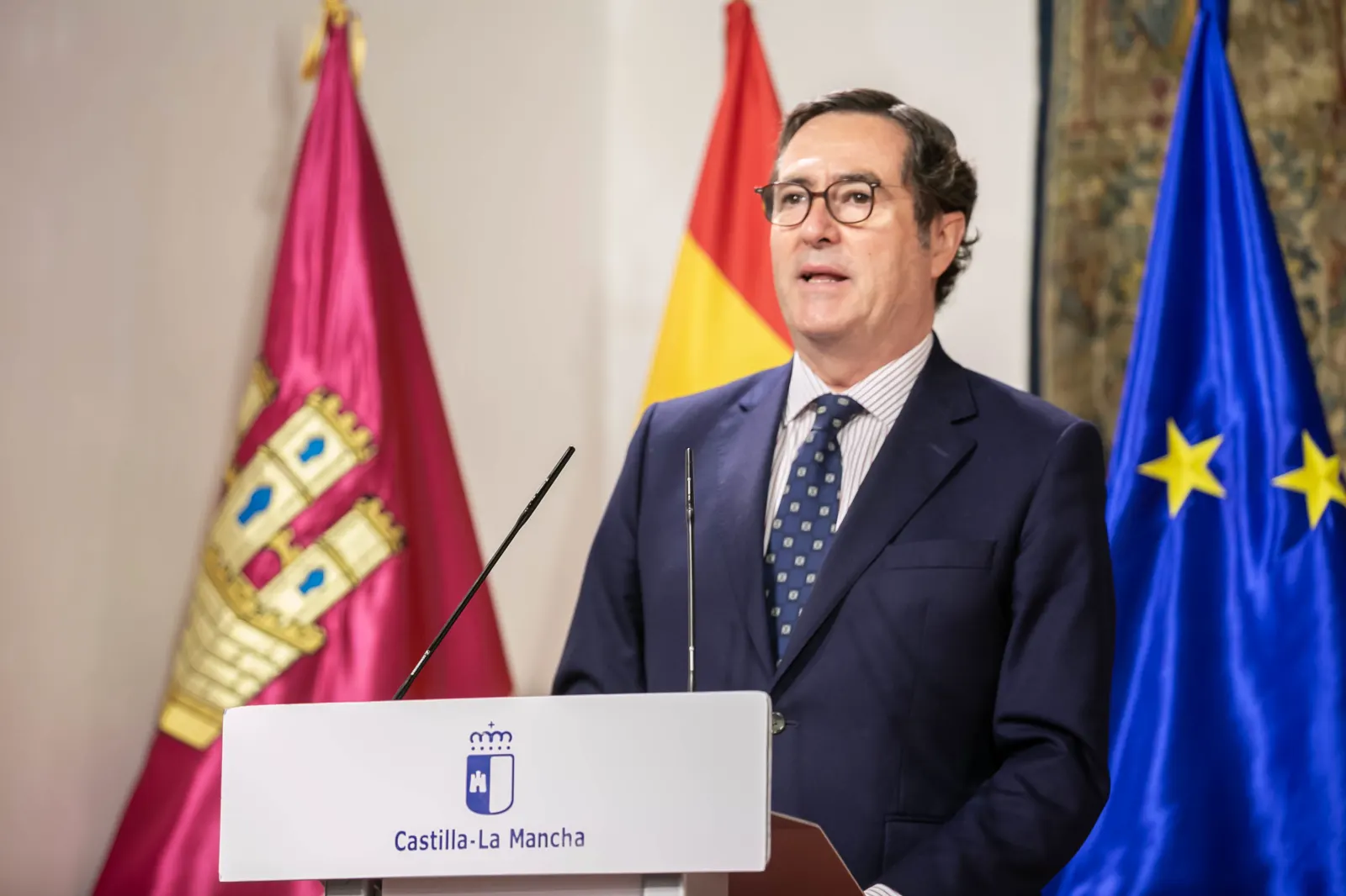 Antonio Garamendi, con traje oscuro, gafas y corbata azul, habla ante un atril con el escudo de Castilla-La Mancha, entre las banderas regional, española y de la Unión Europea.