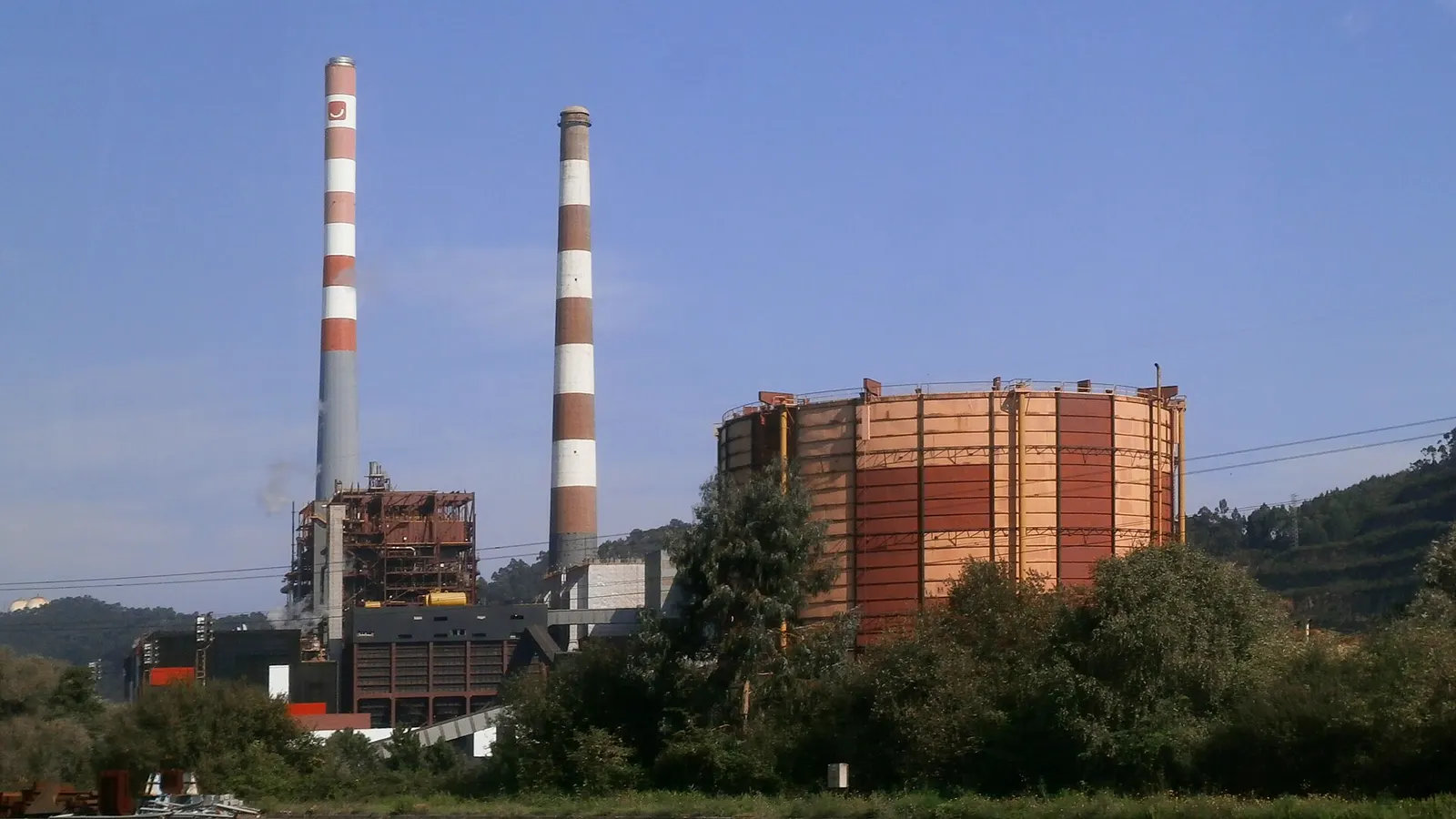 Dos chimeneas de franjas blancas y rojas de una central térmica junto a un gasómetro cilíndrico oxidado, rodeados de vegetación y con el mar al fondo